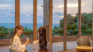 Dos personas sentadas en una terraza con vista al mar, una mesa con un plato y una copa, y una silla.