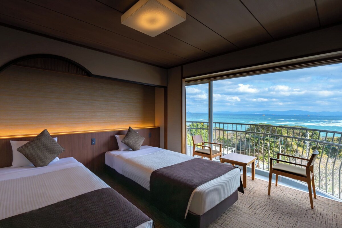 Habitación de hotel con dos camas, un balcón con vista al océano y una pequeña mesa con una silla.