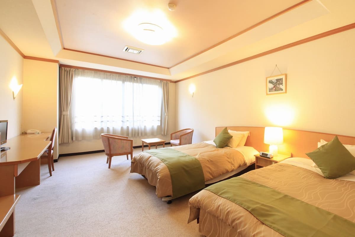 A hotel room with two beds, a desk, a chair, a lamp, and a window with curtains.