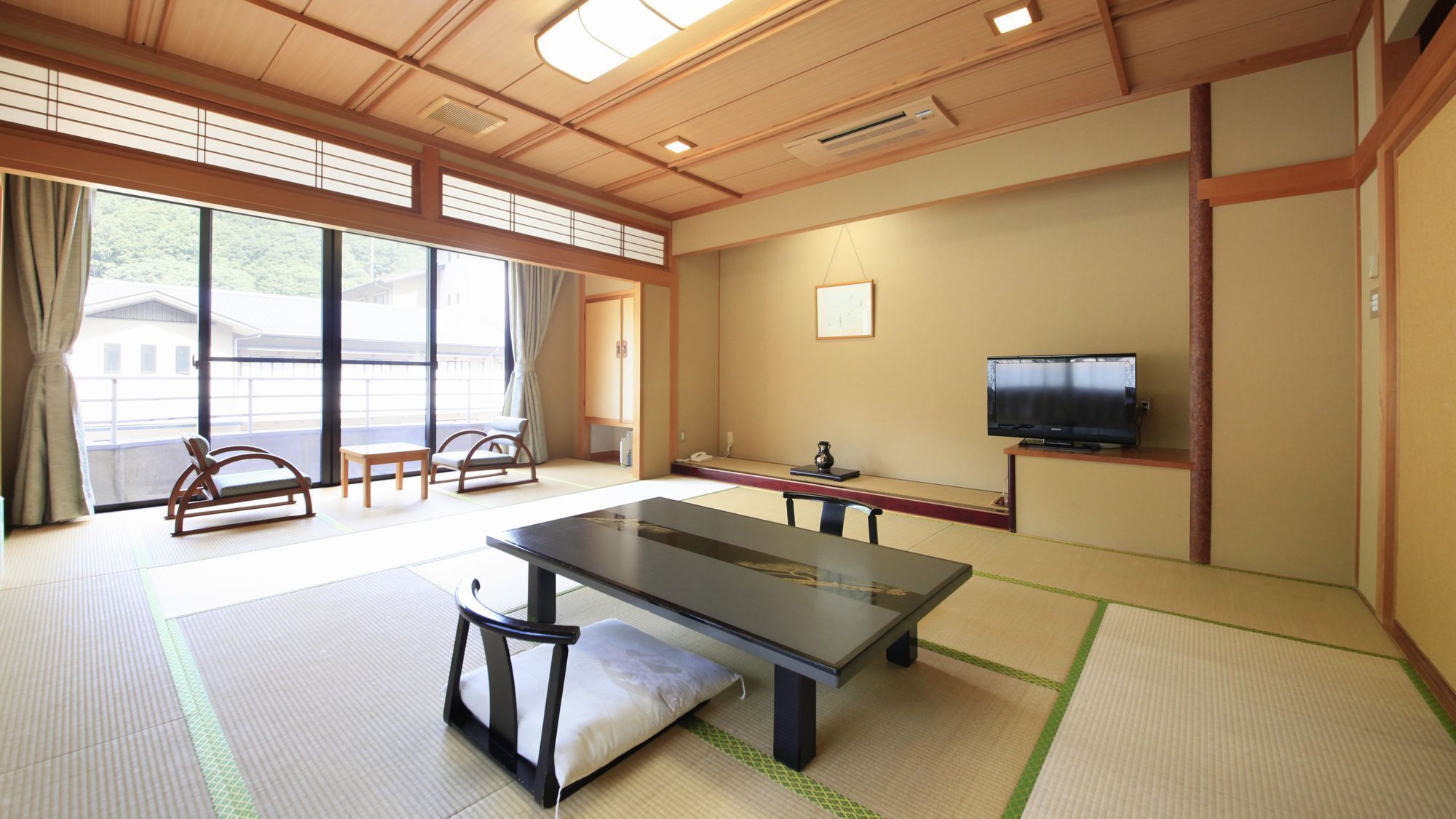 A tatami mat room with a tatami mat table, chairs, a television, and a window with a view of buildings.