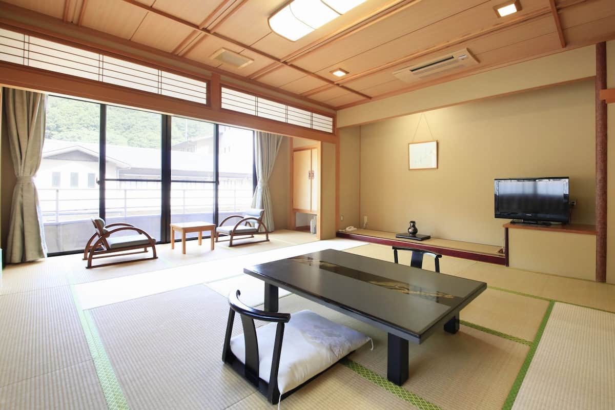 A tatami mat room with a tatami mat table, chairs, a television, and a window with a view of buildings.