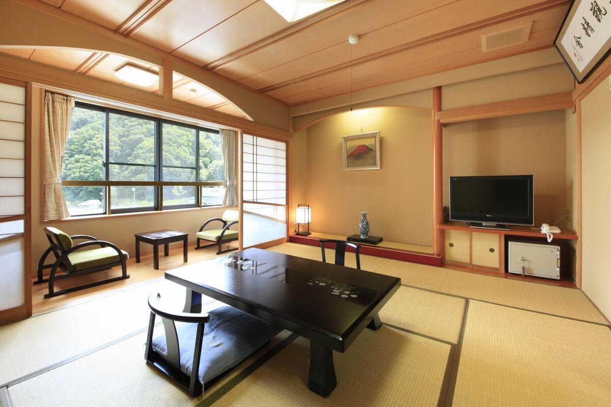 A traditional Japanese room with tatami flooring, a low table, a sofa, a TV, and a window with a view of greenery.