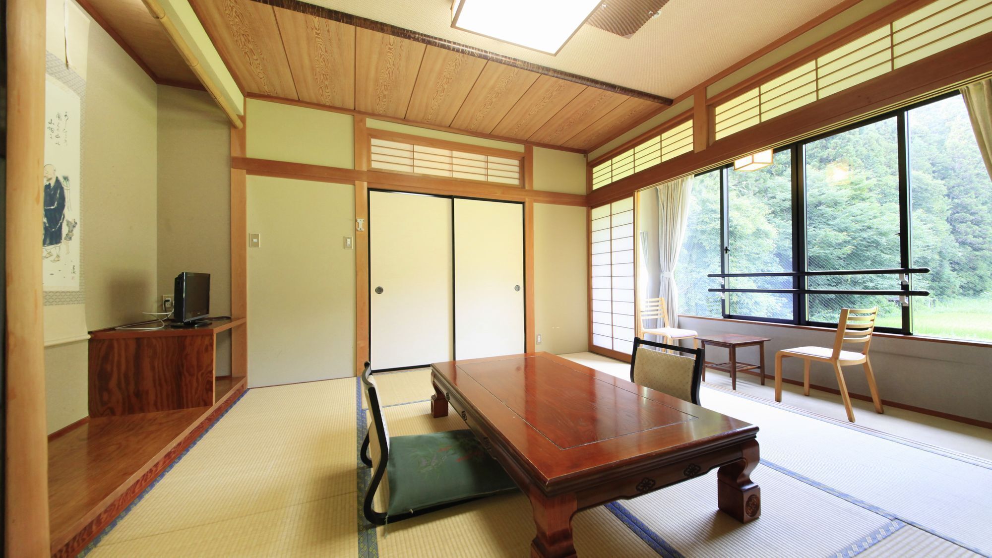 Una habitación japonesa tradicional con suelo de tatami, una mesa baja, sillas, puertas correderas y un ventanal con vistas a zonas verdes.