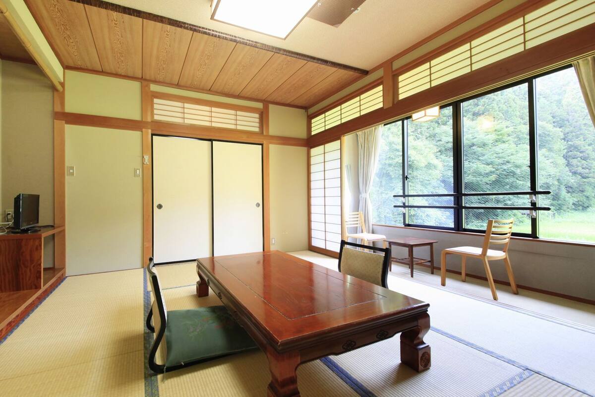 A traditional Japanese room with tatami flooring, a low table, chairs, sliding doors, and a large window with a view of greenery.