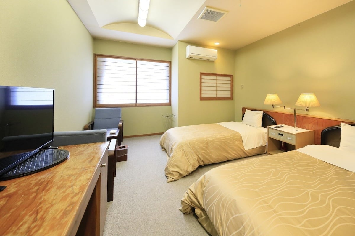 A hotel room with two beds, a desk with a computer, a chair, and a window with blinds.