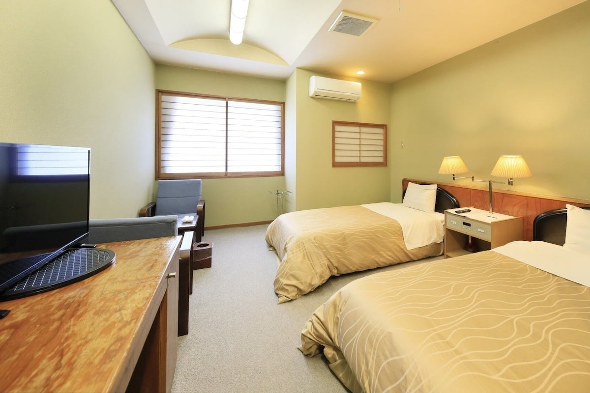 A hotel room with two beds, a desk with a computer, a chair, and a window with blinds.