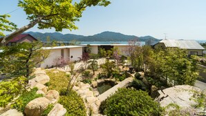 Un jardín japonés tradicional con un estanque de carpas, rodeado de rocas y diversas plantas. Al fondo, se observa un lago y montañas.
