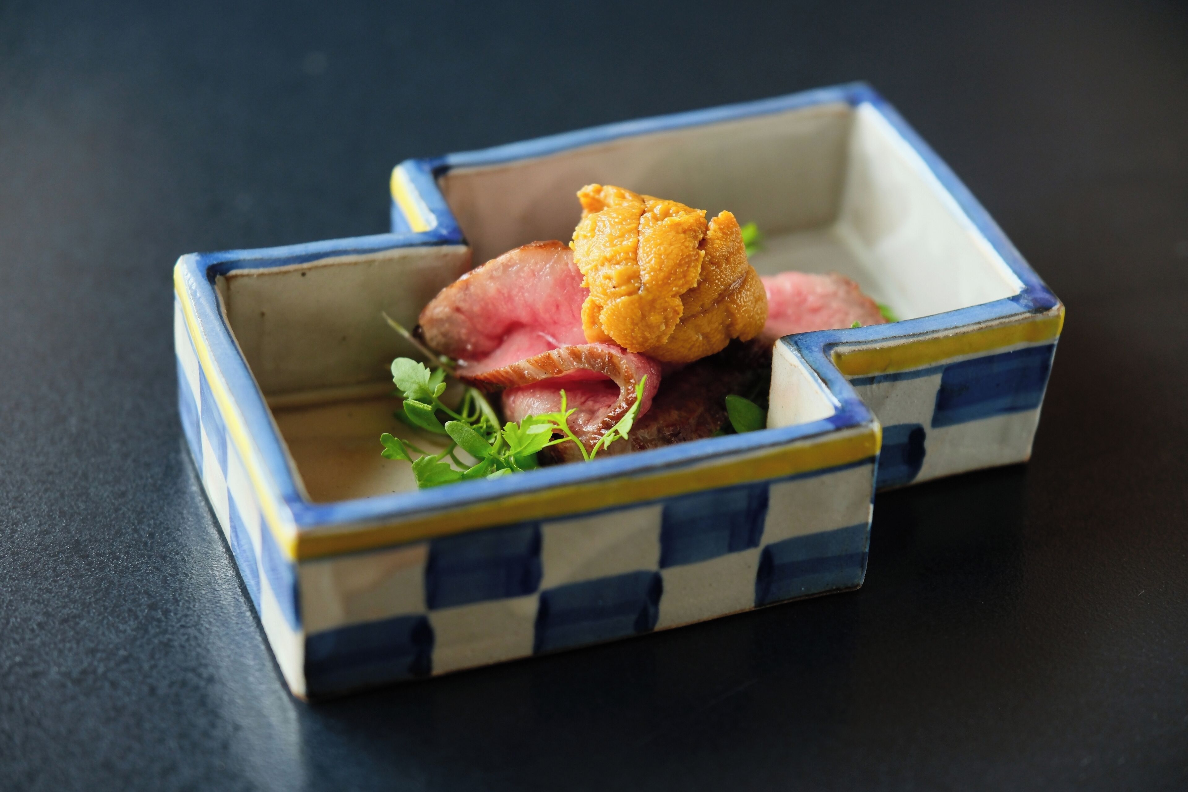 Una pequeña vasija cuadrada de cerámica con diseño de cuadros que contiene rebanadas de carne y un trozo de comida frita, decorada con hierbas, sobre una superficie oscura.