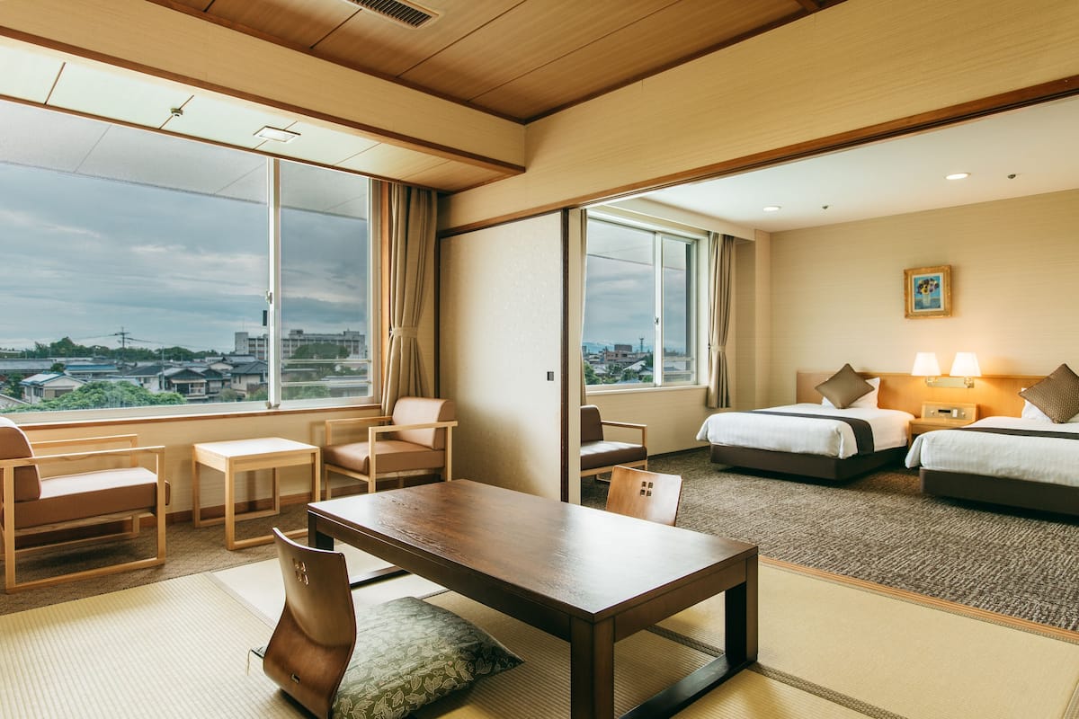 A hotel room with a large window, a wooden table, two beds, and a view of the city.