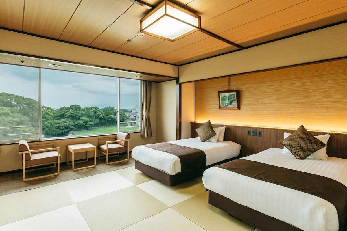 A hotel room with two beds, a sitting area with chairs, a large window with a view of greenery, and a wooden headboard.