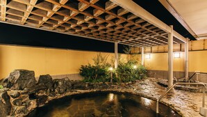 Een stenen warmwaterbron met een houten pergola erboven, omringd door rotsen en planten.