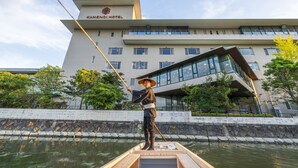 Een persoon in traditionele kledij op een boot voor een hotel met moderne architectuur.