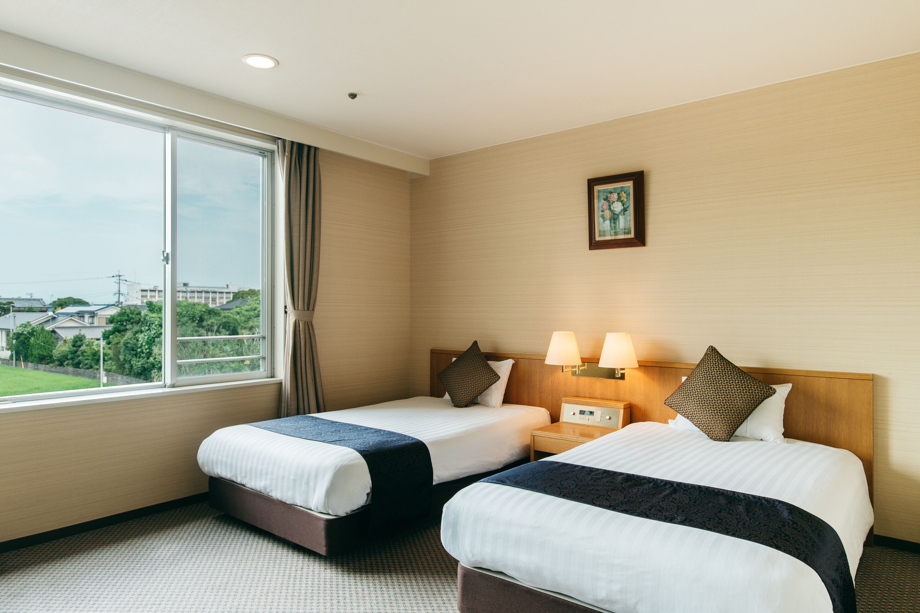 A hotel room with two beds, a large window, a nightstand with lamps, and a framed picture on the wall.