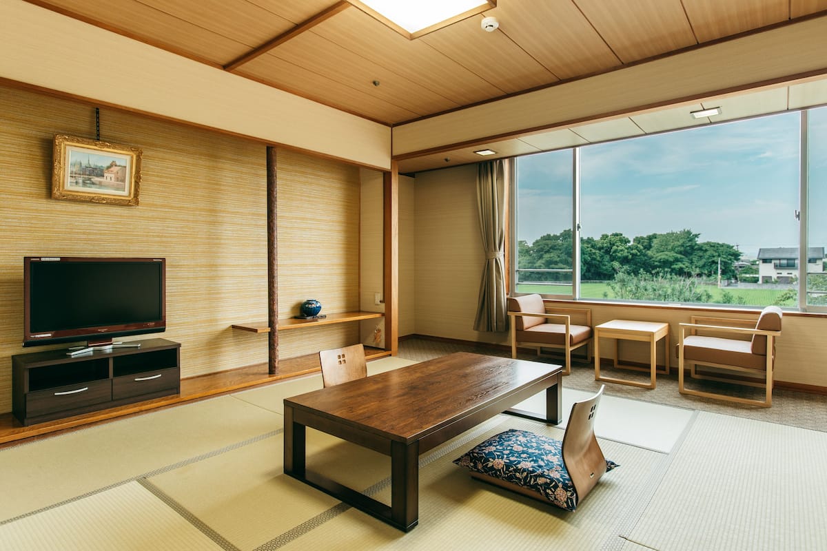 A traditional Japanese-style room with tatami flooring, a low wooden table, a television, and a large window offering a view of greenery and buildings.