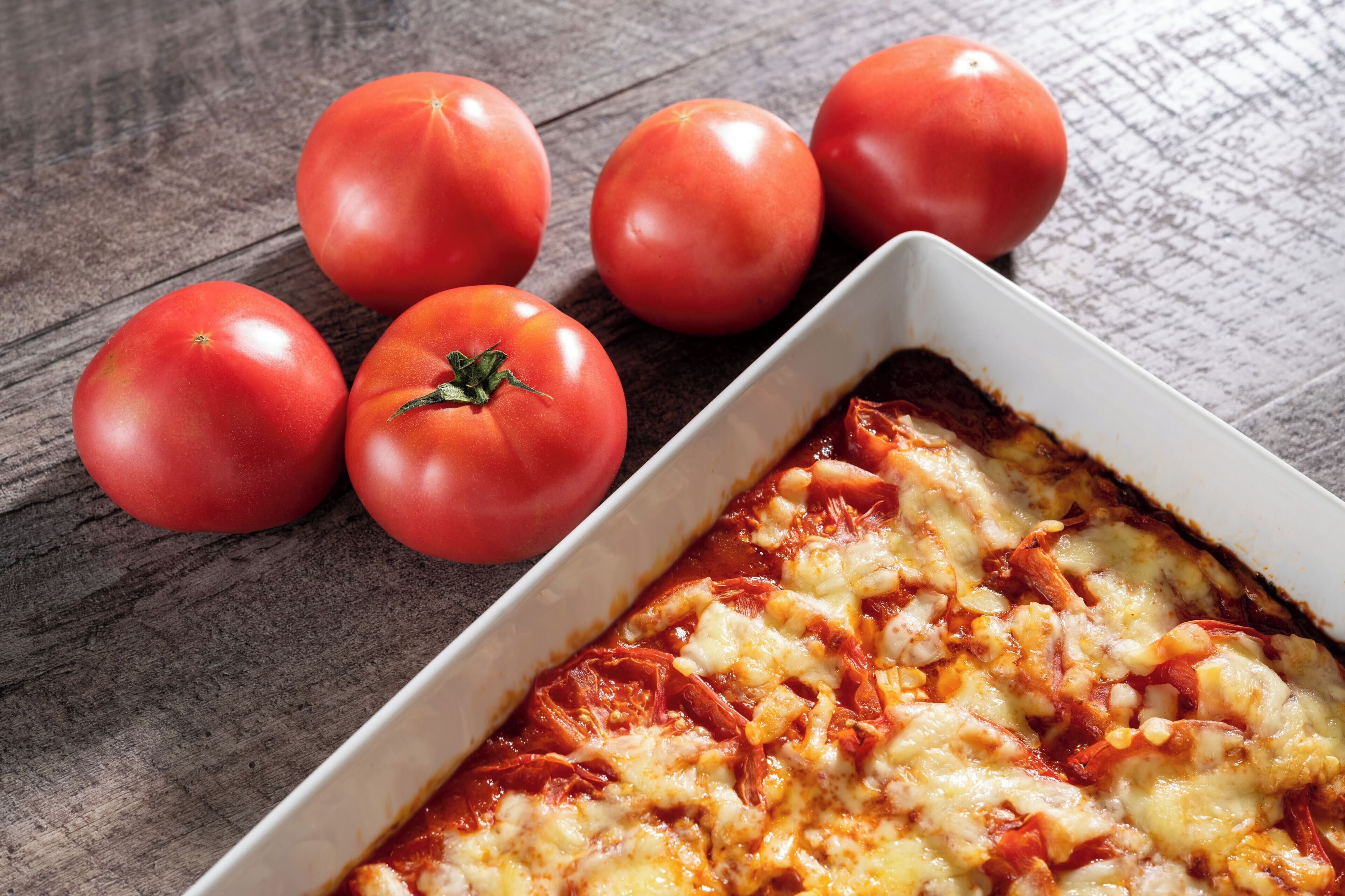 A rectangular baking dish with a baked dish, possibly lasagna, topped with melted cheese and tomato sauce. Five whole tomatoes are placed beside the dish on a wooden surface.