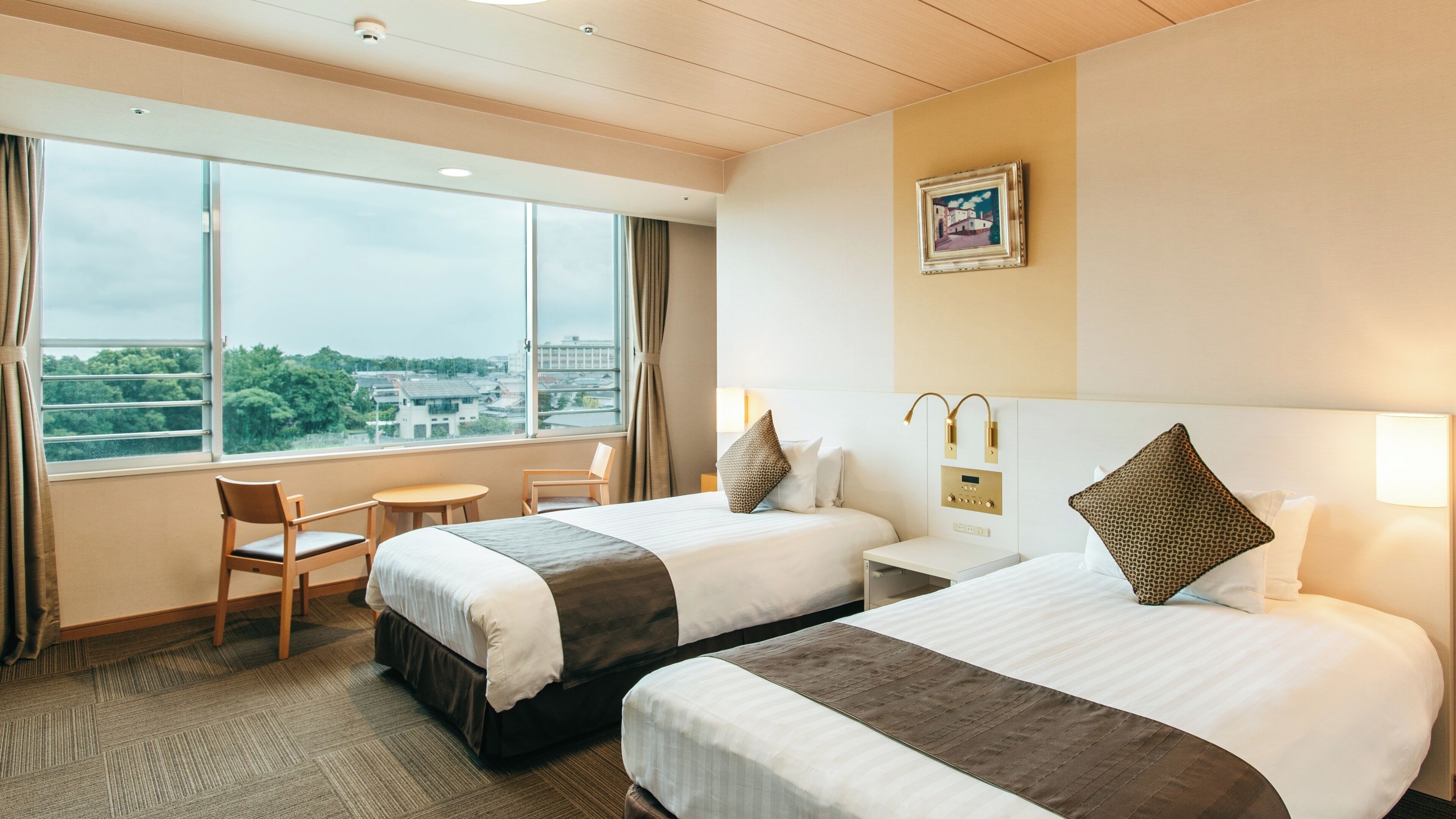 Een hotelkamer met twee bedden, een tafeltje, een stoel, een raam met uitzicht op de stad en een schilderij aan de muur.