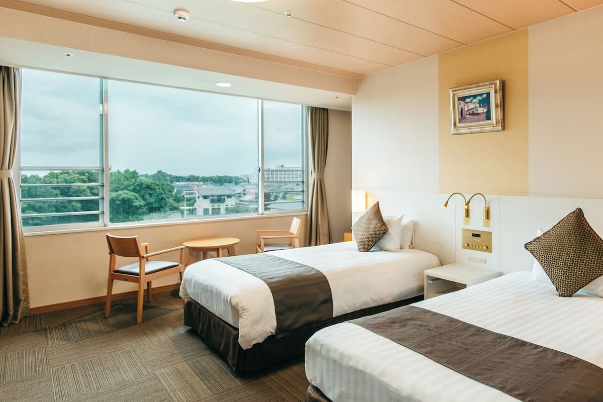 A hotel room with two beds, a small table, a chair, a window with a city view, and a painting on the wall.