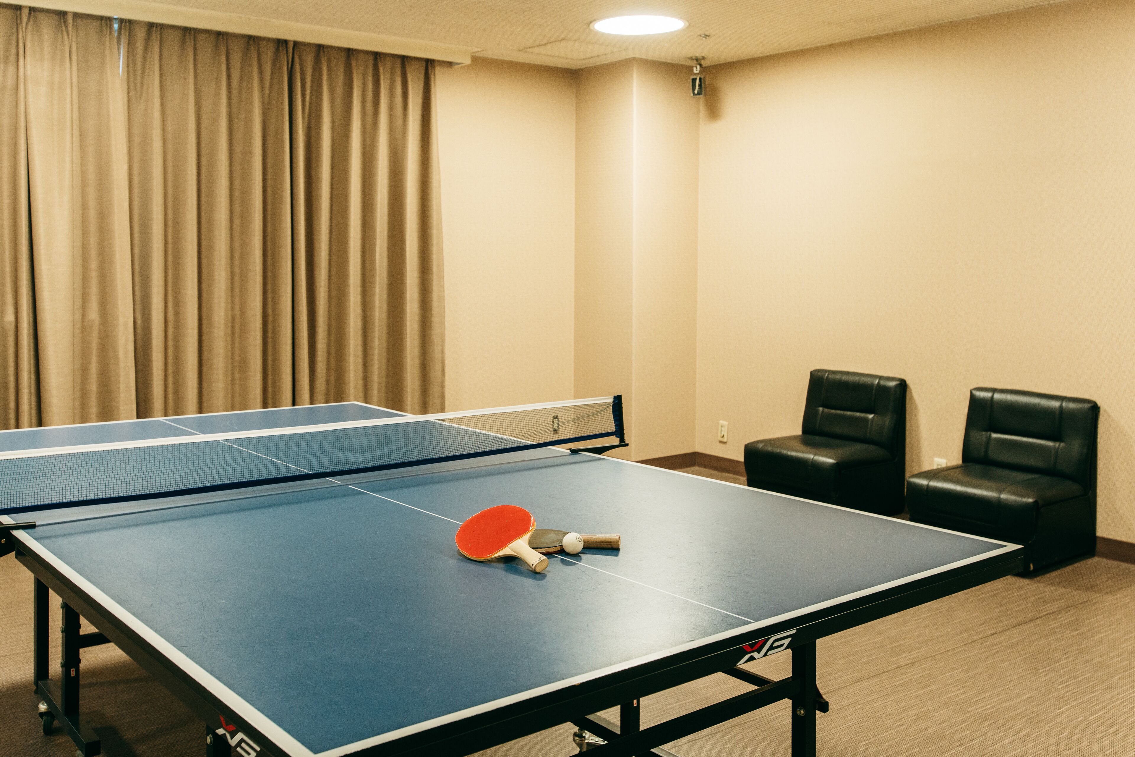 Una mesa de ping pong con una paleta y una pelota de tenis de mesa, dos sillas de cuero negro y una cortina color beige en una habitación con paredes y alfombra color beige.