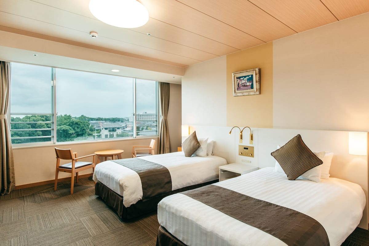 A hotel room with two beds, a desk, and a chair. There is a large window with a view of buildings and trees.