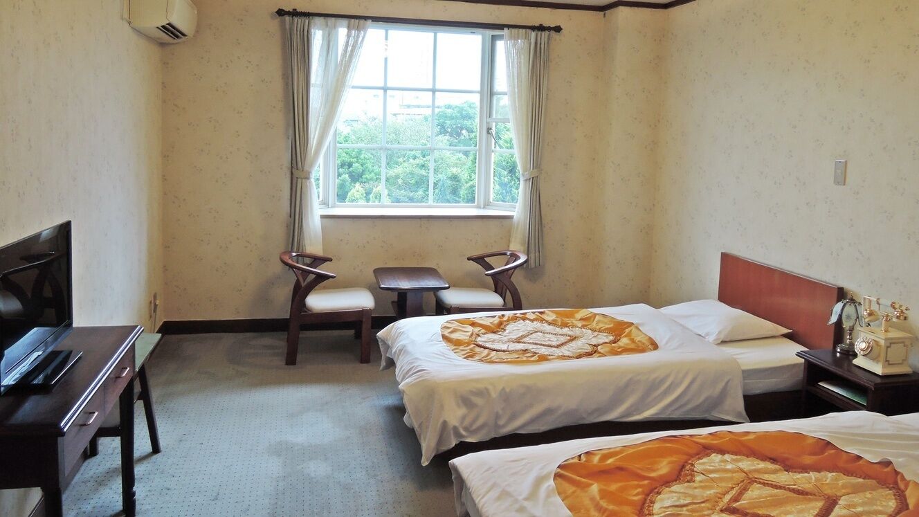 A hotel room with two beds, a desk, a chair, a window with curtains, and a small table.
