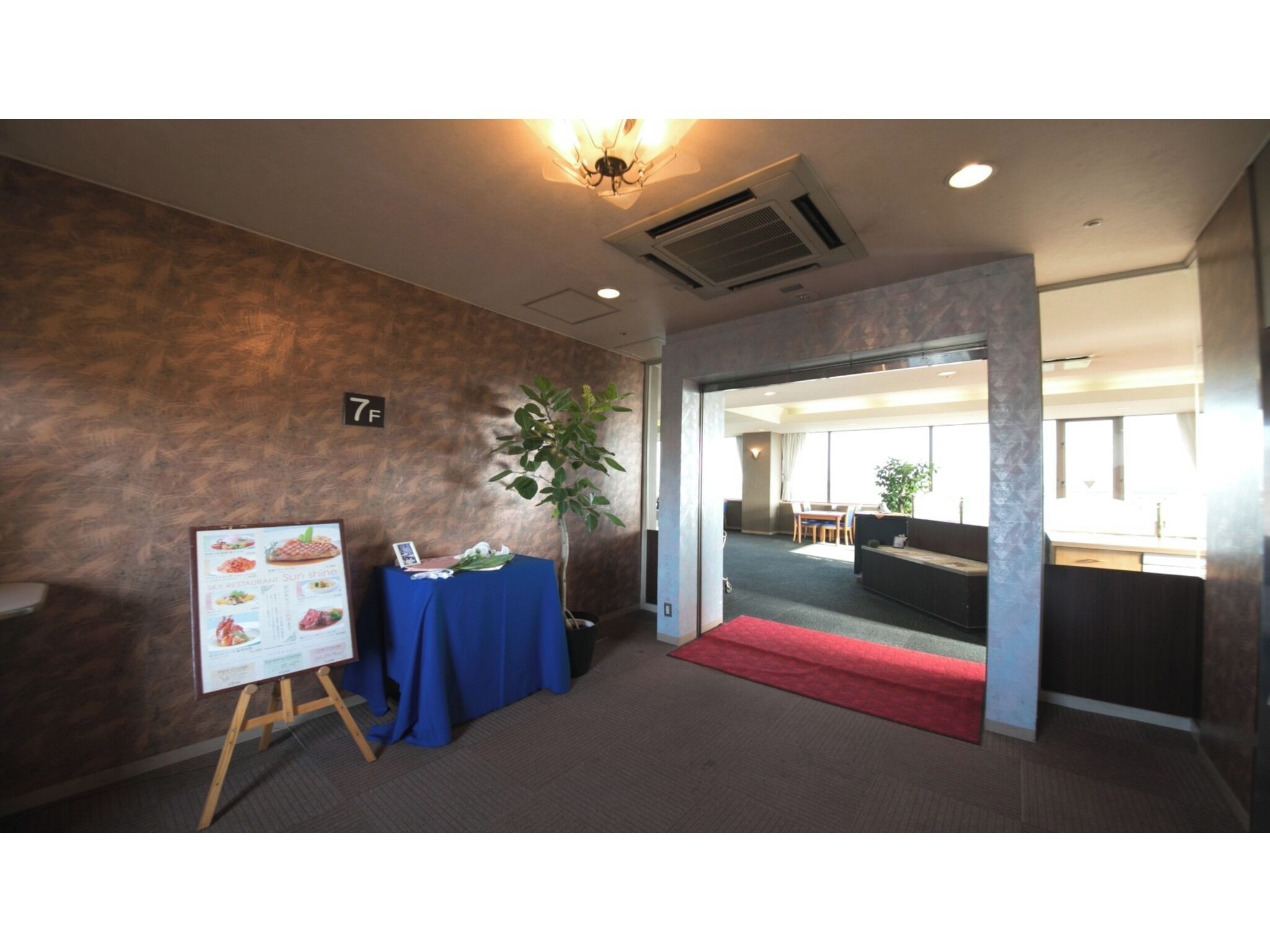 A hotel room with a wooden floor, a red carpet, a table with a blue tablecloth, a plant, and a wall-mounted air conditioning unit.