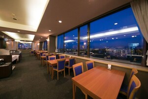 A hotel dining area with tables and chairs, large windows with a city view, and a carpeted floor.