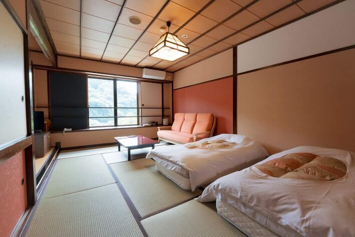 A room with tatami flooring, a sofa, two beds, a small table, and a window with a view.