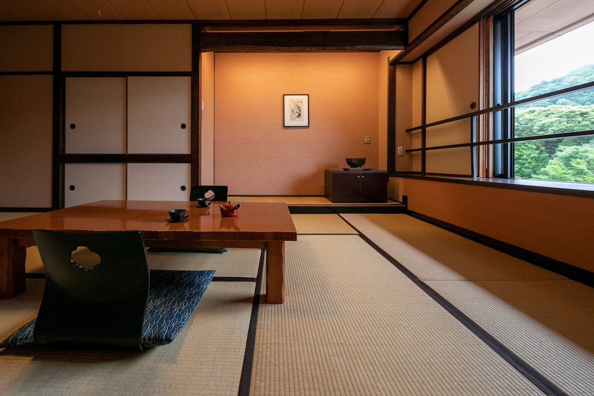 A traditional Japanese room with tatami flooring, a low table, a chair, and a window with a view of greenery.
