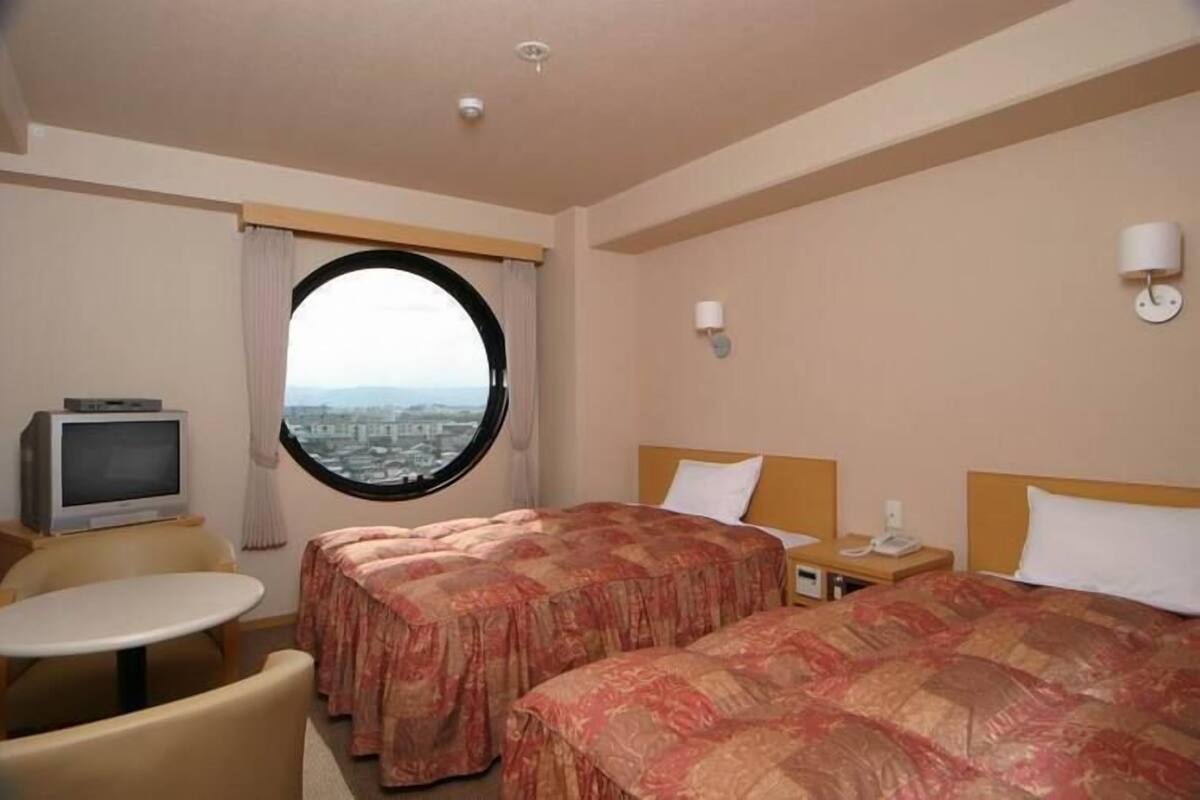 A hotel room with two beds, a TV, a round window, and a view of the city.