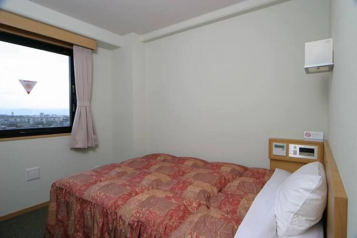 A hotel room with a bed, a window with curtains, a wall-mounted air conditioning unit, and a view of the cityscape.