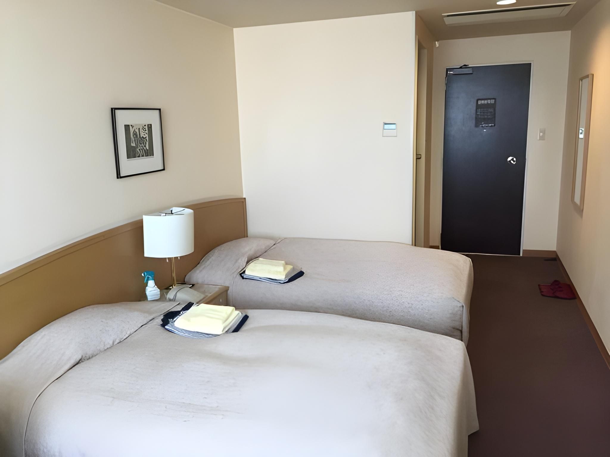 A hotel room with two beds, a bedside table with a lamp, a framed picture on the wall, and a door in the background.