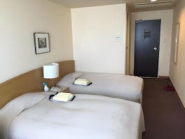 Habitación de hotel con dos camas, una mesita de noche con lámpara, un cuadro enmarcado en la pared y una puerta al fondo.