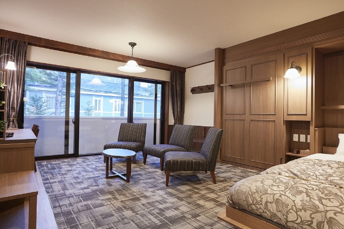 A hotel room with a large bed, two armchairs, a small table, a balcony with curtains, and wooden paneling.