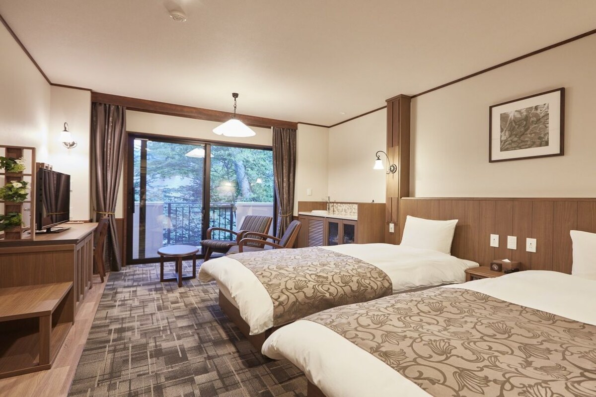 A hotel room with two beds, a desk, a chair, a balcony, and a large window.
