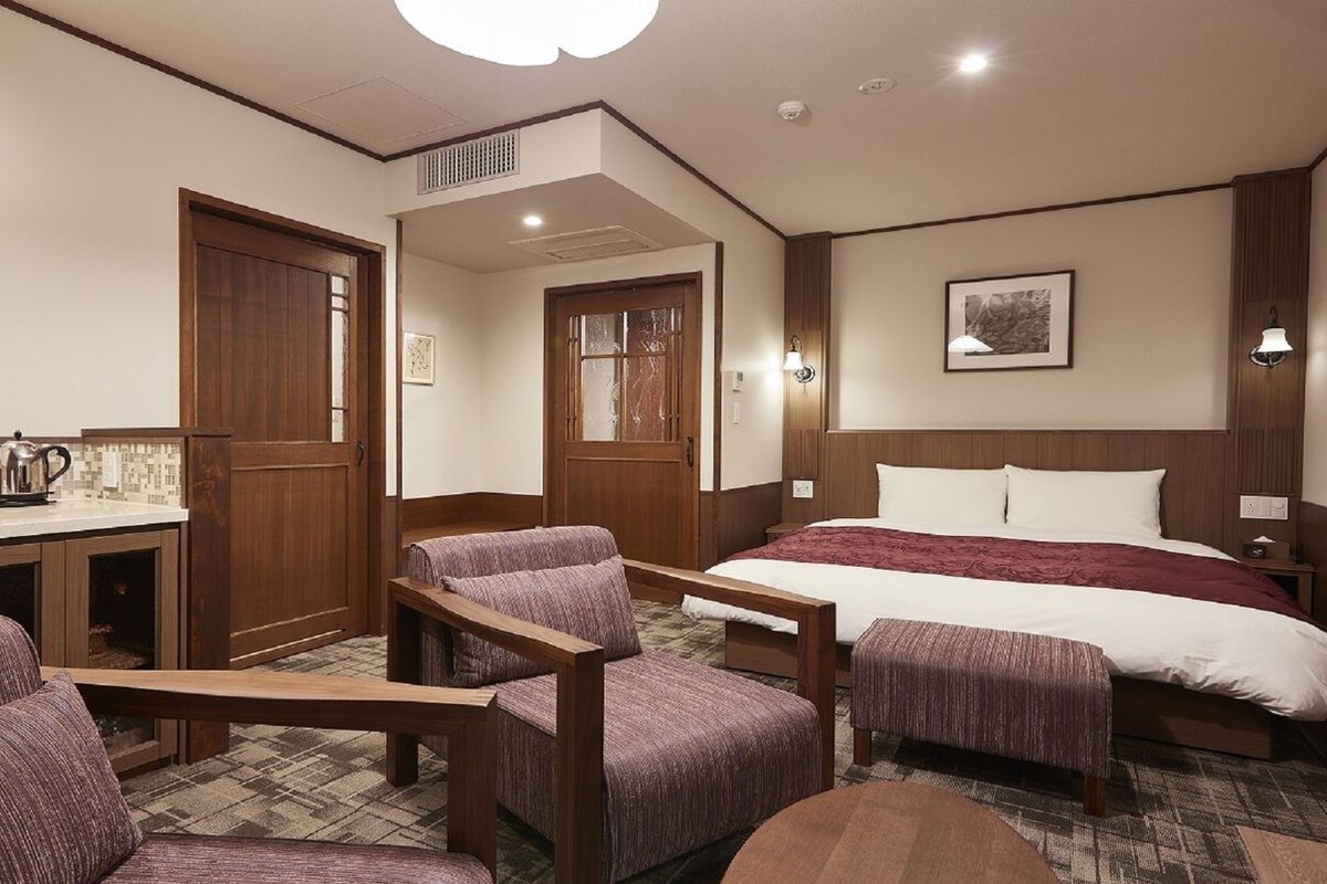 A hotel room with a bed, a desk, two armchairs, and a small table.