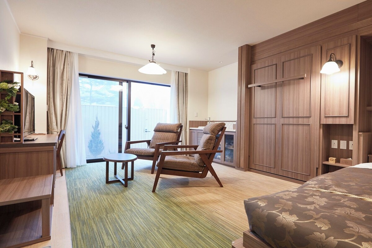 A hotel room with a bed, two armchairs, a desk, a small table, and a view of the outdoors through a sliding door.