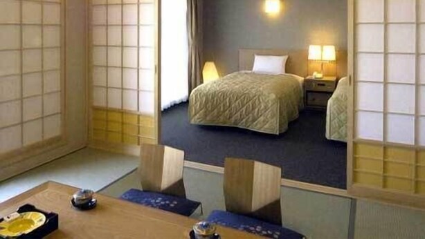 A traditional Japanese-style room with tatami flooring, a bed with a quilted cover, a nightstand with a lamp, and a sliding door with a view of the outside. - Hotel Ayase (Rishifuji)
