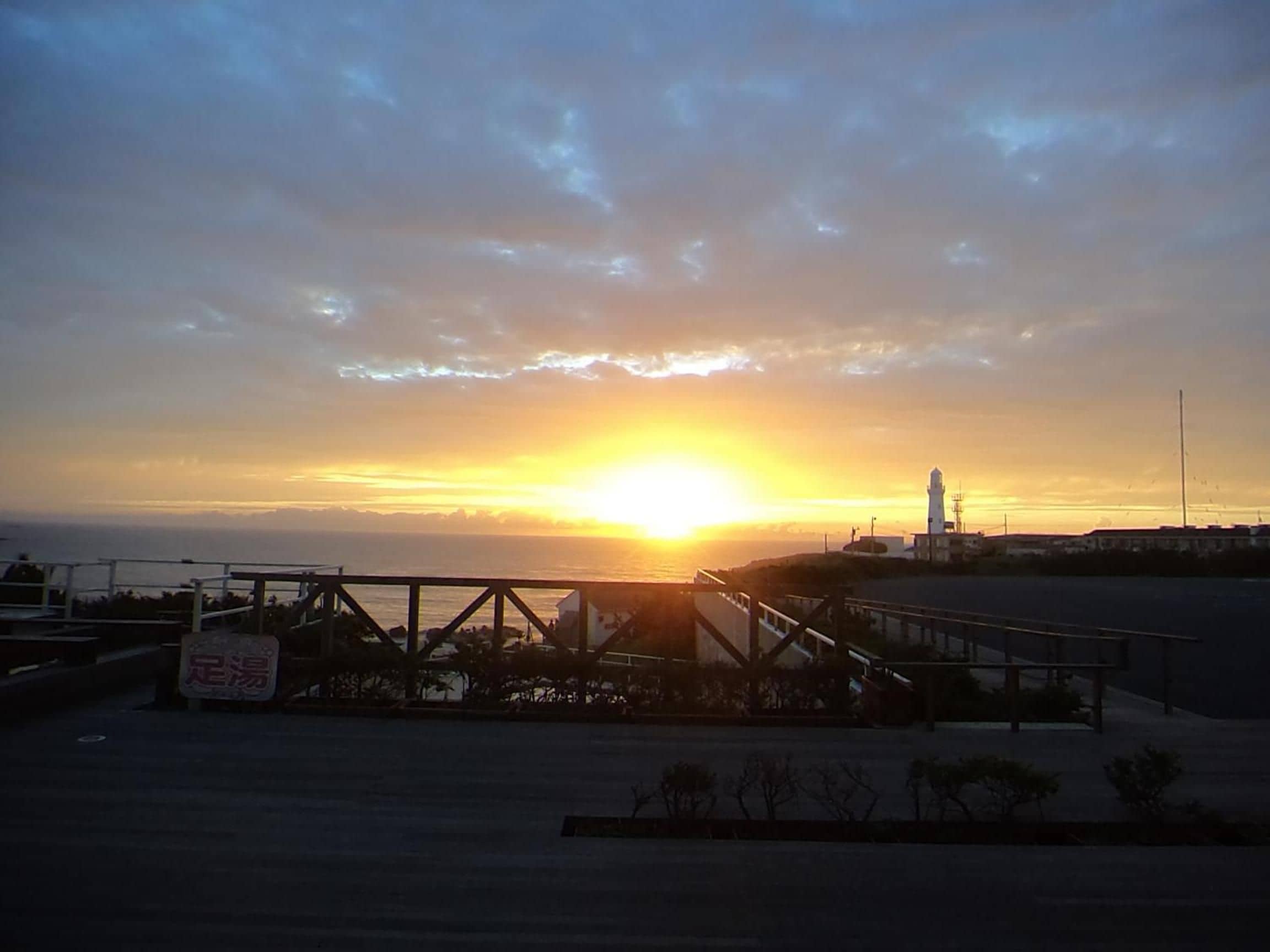 Un coucher de soleil sur une zone côtière, avec un phare et un pont piétonnier.