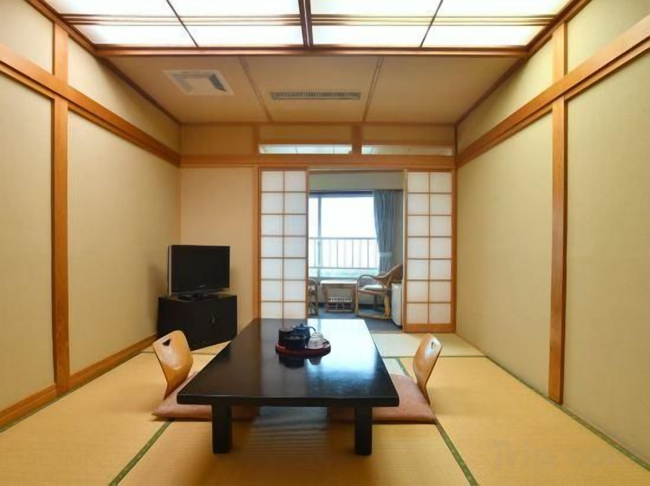Une pièce de style japonais traditionnel, avec un sol recouvert de tatamis, une table basse, des chaises, un téléviseur et une porte coulissante donnant sur un balcon.