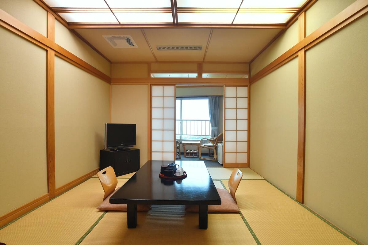 A traditional Japanese room with tatami flooring, a low table, a television, and sliding doors leading to another room.