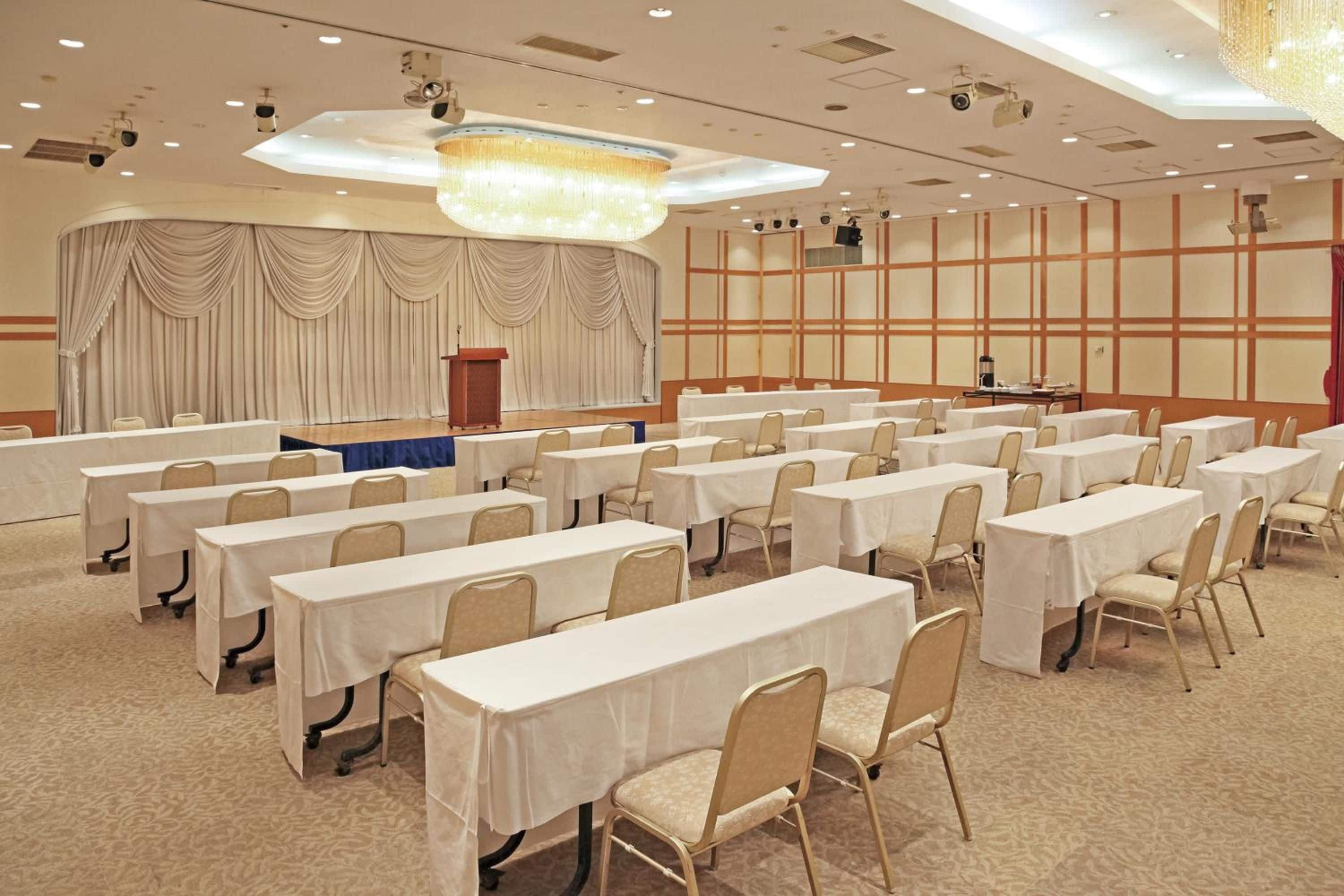 A conference room with rows of tables and chairs, a stage with curtains, and a chandelier.