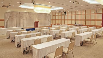 A conference room with rows of tables and chairs, a stage with curtains, and a chandelier.