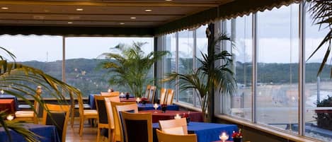 A restaurant with tables and chairs, large windows, and indoor plants.