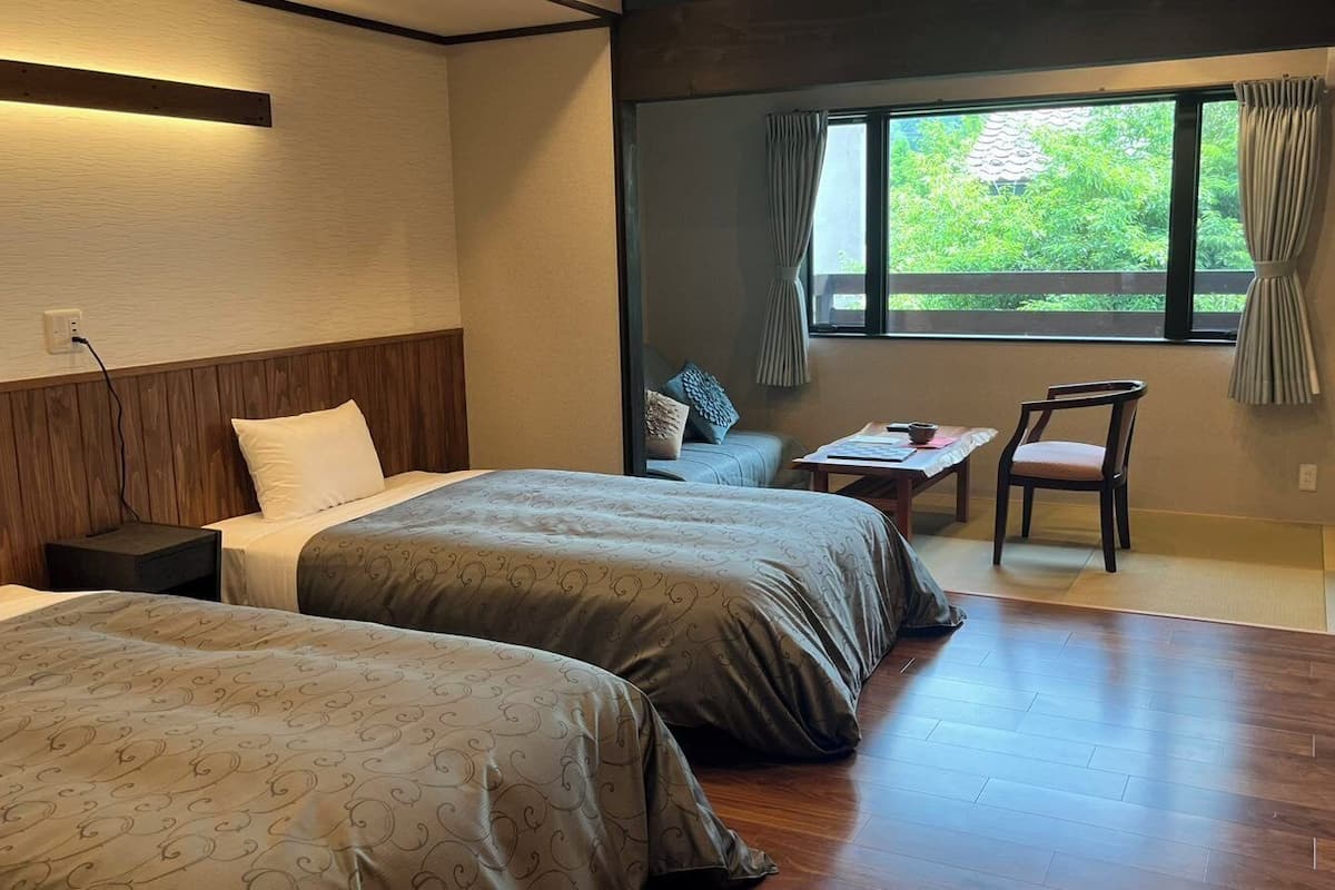 A hotel room with two beds, a small table, a chair, a window with curtains, and a wooden floor.