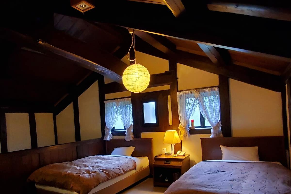 A room with two beds, a hanging light fixture, a bedside table with a lamp, and a window with curtains.