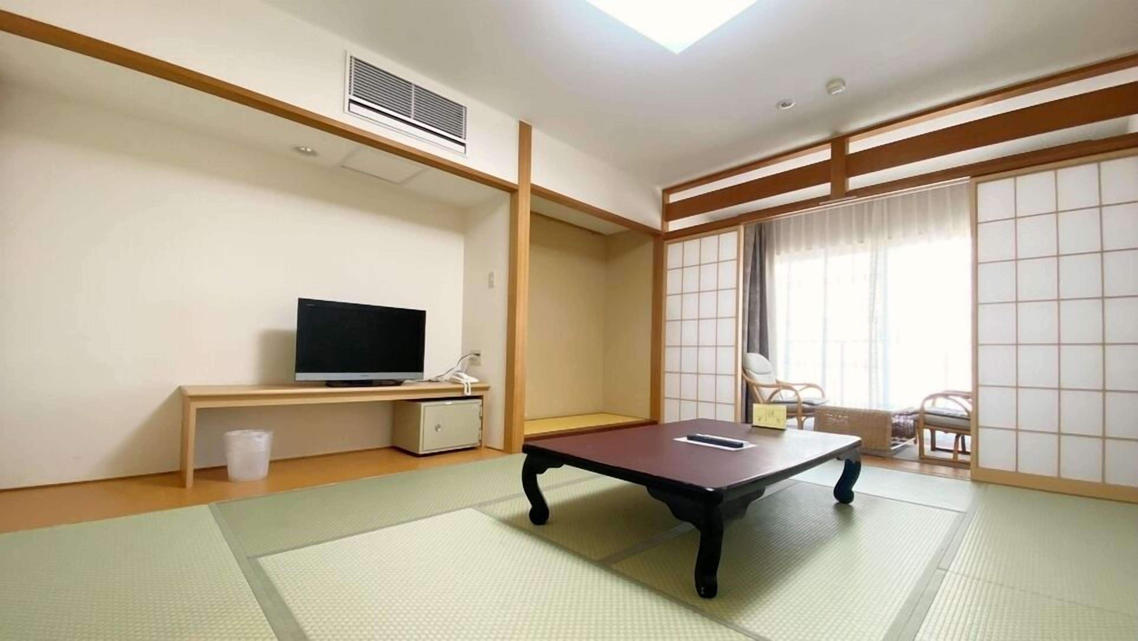 Ein Zimmer mit Tatami-Boden, einem Fernseher auf einem niedrigen Beistelltisch, einem Schreibtisch, einem Stuhl und einer Schiebetür, die auf einen Balkon führt.