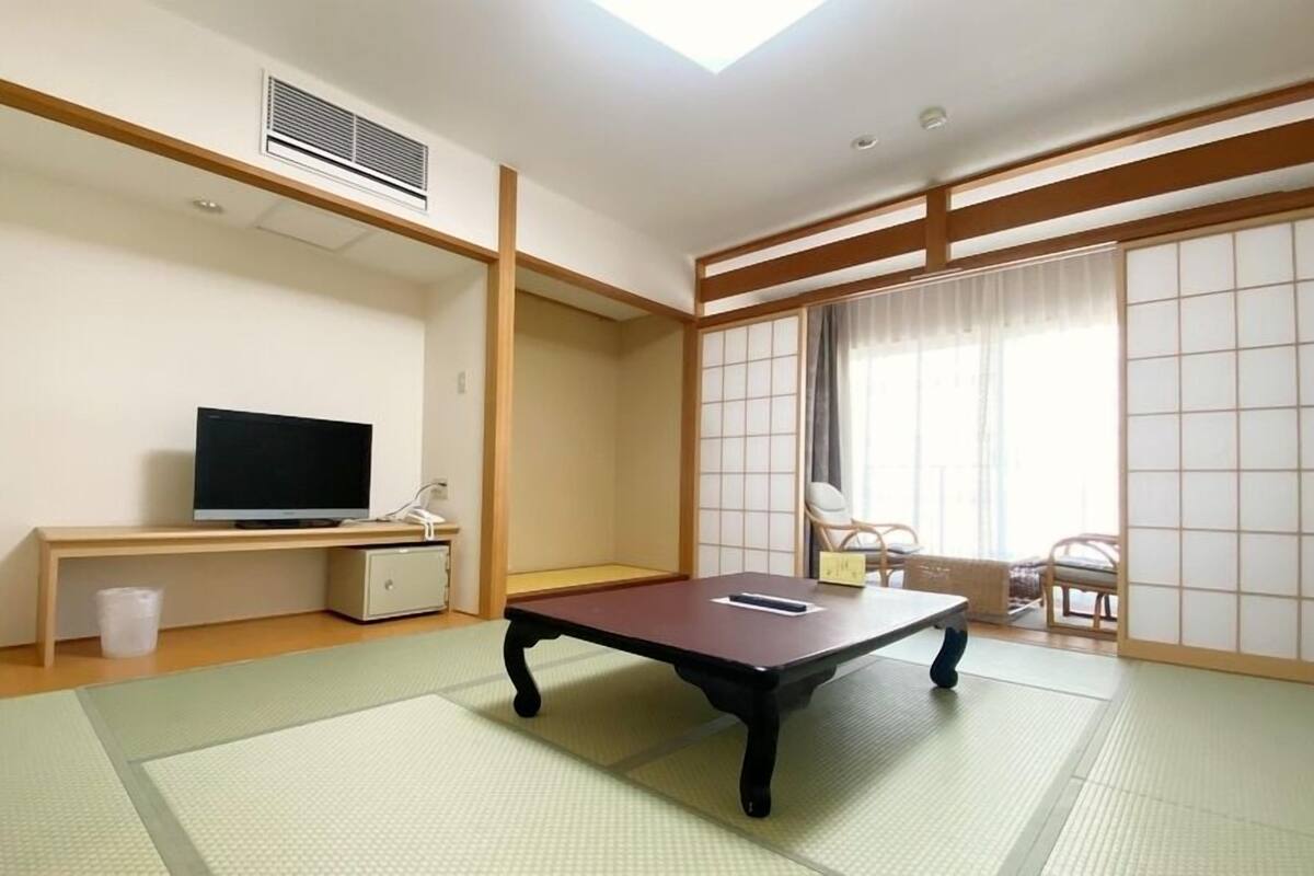 Una habitación con suelo de tatami, una televisión sobre un mueble bajo, un escritorio, una silla y una puerta corredera que da a un balcón.