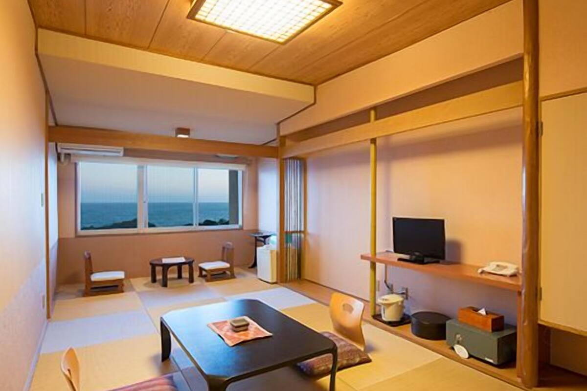 Ein traditioneller japanischer Raum mit Tatami-Böden, einem niedrigen Tisch, einem Fernseher auf einem Regal und Meerblick durch das Fenster.