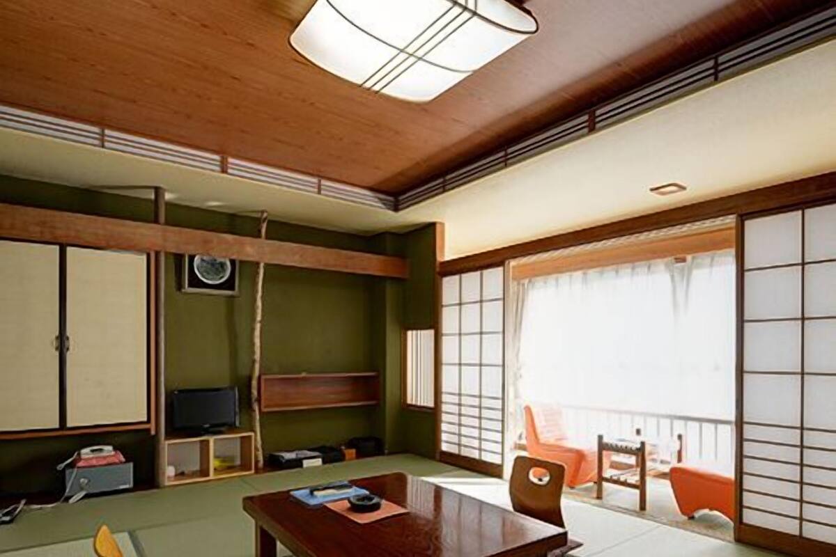 Ein traditioneller japanischer Raum mit Tatami-Bodenbelag, einem niedrigen Holztisch, einem Fernseher und Schiebetüren.