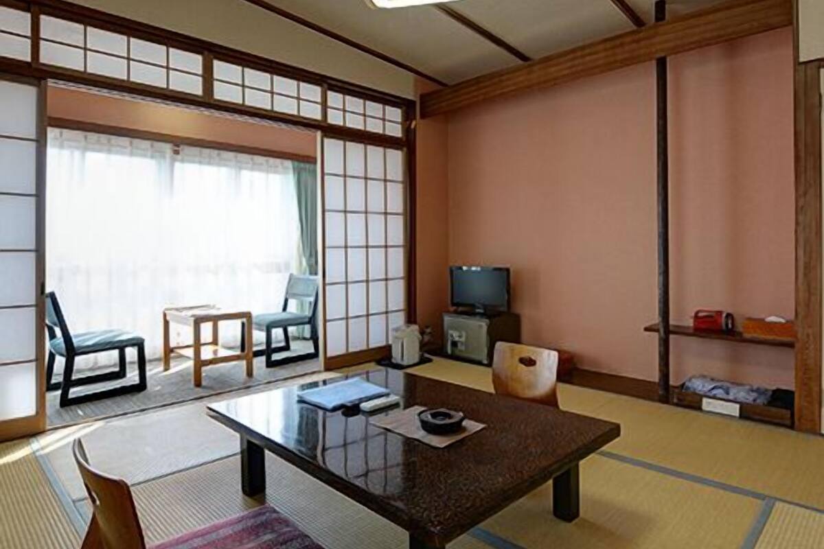 Ein traditioneller japanischer Raum mit Tatami-Boden, einem niedrigen Tisch, Stühlen, einem Fernseher und Schiebetüren.